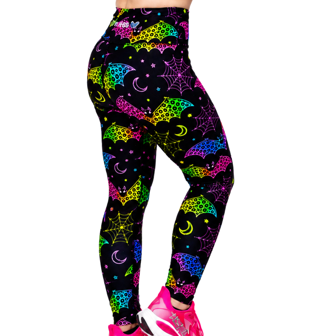 A person wearing high-waisted leggings with a neon multicolour bat and spiderweb print on a black background, paired with bright pink running shoes, shown against a plain white studio background.