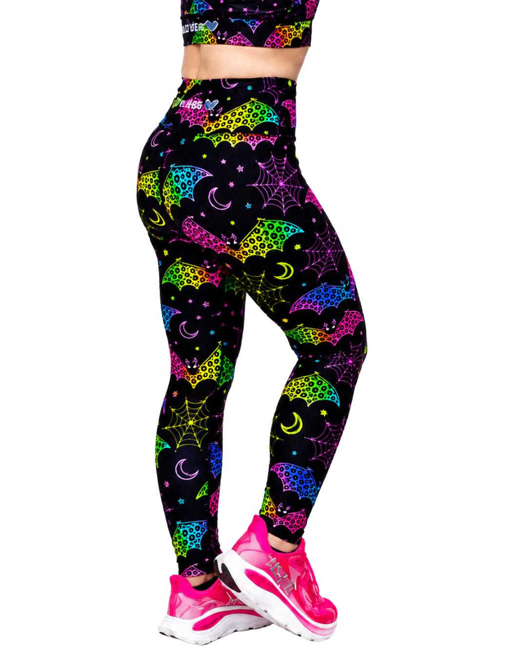 A person wearing high-waisted leggings with a neon multicolour bat and spiderweb print on a black background, paired with bright pink running shoes, shown against a plain white studio background.