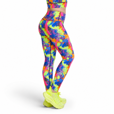 A person shown from the waist down wearing high-waisted leggings with a bright multicolour paint splash pattern in neon pink, yellow, green, blue, and purple, paired with neon yellow sneakers, against a plain white background.