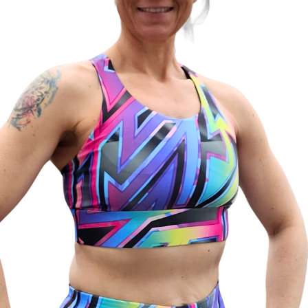 A close-up of a woman wearing a colorful athletic sports bra with a bold geometric pattern in pink, blue, purple, and green, paired with matching athletic bottoms against a plain white background.